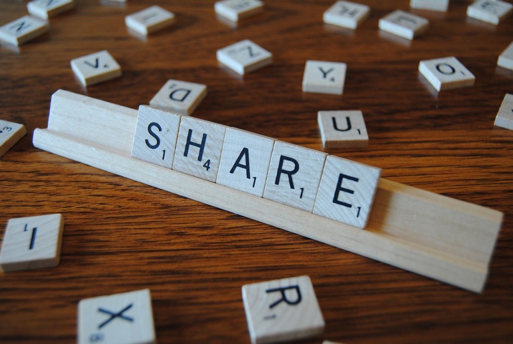 Scrabble tiles spelling "SHARE" on a wooden tile rack, with scattered tiles in the background.